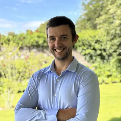 Man in light blue shirt standing with arms crossed in a sunny garden.