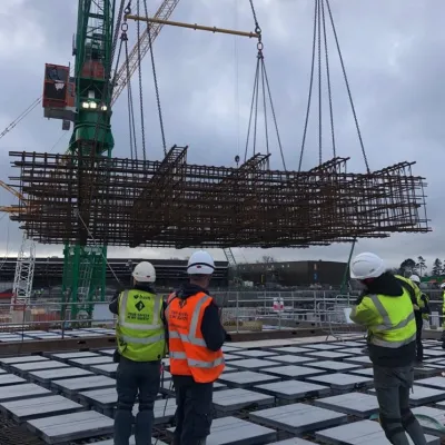 Construction workers in safety gear observing a large metal framework being lifted by a crane at a building site.