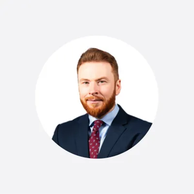 Professional headshot of a red-bearded man in a navy suit and red tie, circular crop.