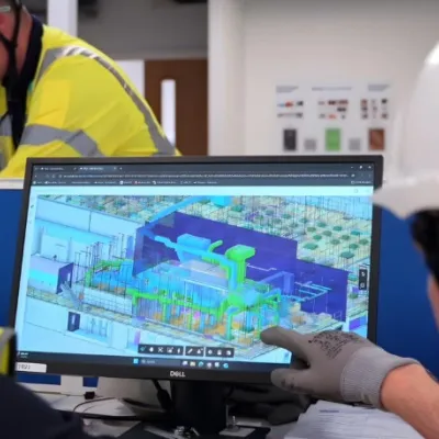 Two workers in high-visibility jackets and hard hats reviewing data on a computer monitor.