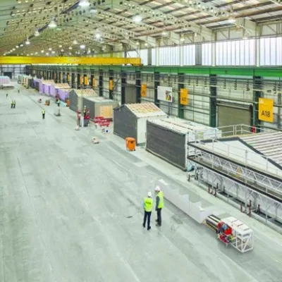 Large industrial warehouse interior with workers in high-visibility vests walking along a wide concrete floor.