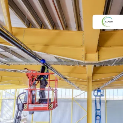 Worker on a red aerial lift working on yellow industrial ceiling beams indoors.
