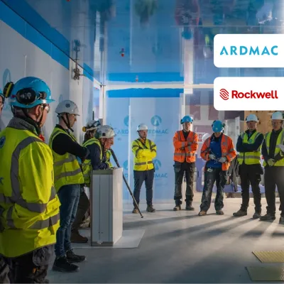 Group of construction workers in high-visibility vests and hard hats at an indoor briefing, Ardmac and Rockwell logos.