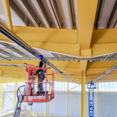 Worker on a yellow aerial lift platform installing ceiling components in an industrial building.