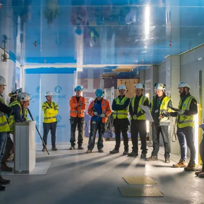 Group of construction workers in hard hats and high-visibility vests gathered in an industrial facility.