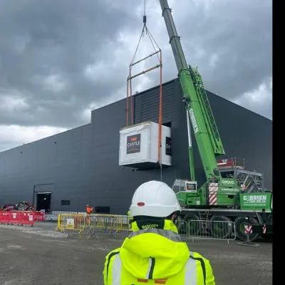 Worker in high-vis jacket watches a crane lifting equipment at a large industrial building.