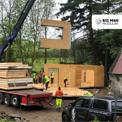 Workers installing a large modular wall panel via crane onto a construction site surrounded by trees.