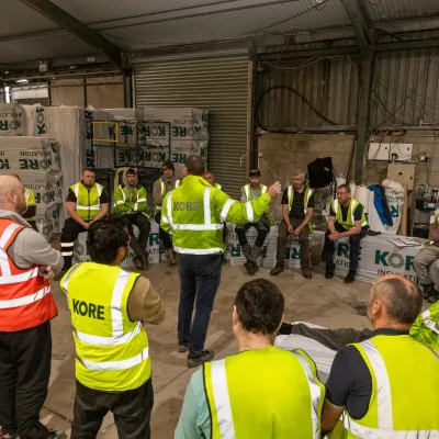 Group of workers in yellow safety vests gathered inside a warehouse for a briefing.