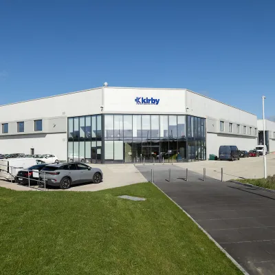 Modern industrial building with "Eleby" signage, cars parked outside under a cloudy sky.