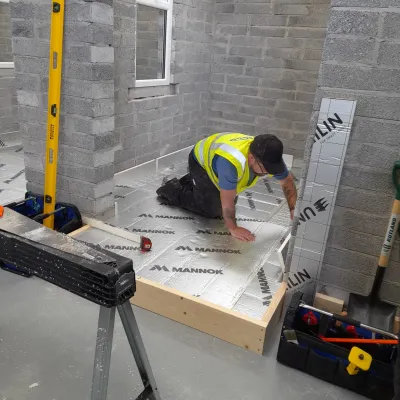 Worker in yellow safety vest laying flooring in a concrete block room under construction.