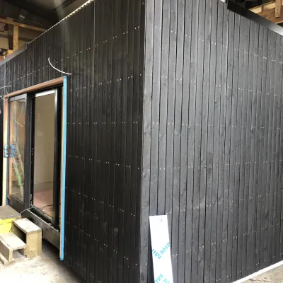 Small modular building with dark charred wood siding and sliding glass door, under construction indoors.