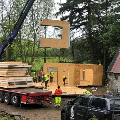 A crane lifts a large wooden frame module onto a modular home construction site surrounded by trees.