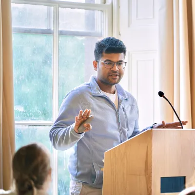 Young man with glasses speaking at a wooden podium in an elegant room with audience.