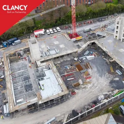 Aerial view of an active construction site with foundations, a red crane, and surrounding buildings.