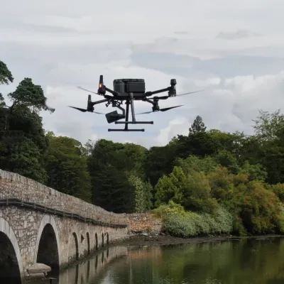 Black drone flying over a stone arch bridge above calm river, surrounded by lush green trees.