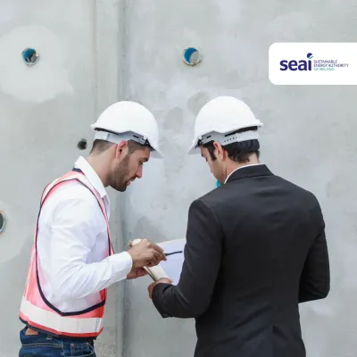 Two construction workers in white hard hats reviewing documents at a concrete wall, SEAI logo visible.