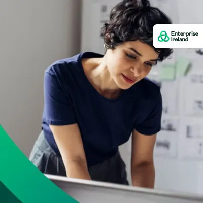 Woman in navy top leaning over a laptop, smiling; Enterprise Ireland logo in top right corner.