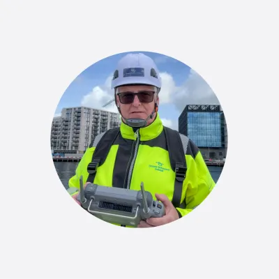 Person in neon yellow safety jacket and white hard hat holding equipment outdoors near buildings.