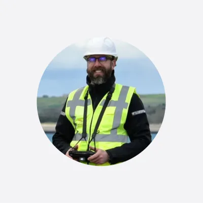 Bearded man wearing a white hard hat and yellow high-visibility vest outdoors near water.