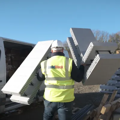 Worker in yellow safety vest carrying large white panels near a white van outdoors.