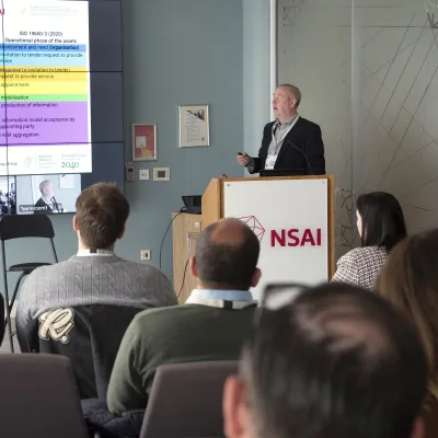 Man presenting at an NSAI conference, standing at a podium beside a colorful data slide.