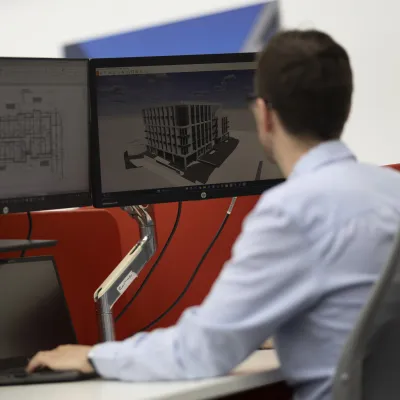 Man in blue shirt working at dual monitor workstation displaying CAD designs.