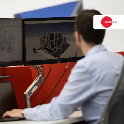 Man working at dual monitors displaying 3D CAD building model in an office setting.