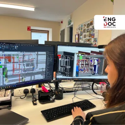 Woman working at dual monitors displaying technical CAD engineering drawings in an office.