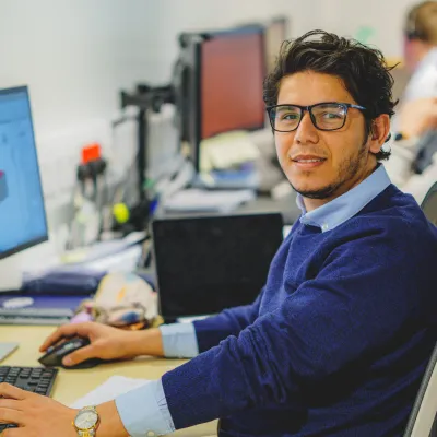 Young man in a blue suit smiling at his desk with a 3D CAD model on his computer screen.