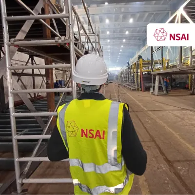 Person in NSAI high-visibility vest and hard hat inspecting a large industrial construction site.