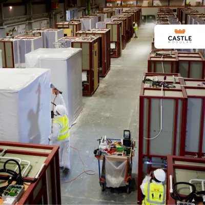 Factory workers preparing prefabricated construction modules inside a large manufacturing facility. Numerous partially assembled modules are arranged throughout the factory floor.