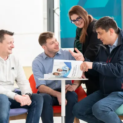 A picture of a group of individuals sitting around a small circular desk looking through a Kirby booklet and one of the man is wearing a coat that has the Kirby Academy branding