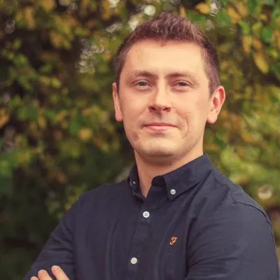 Smiling man in a dark shirt with arms crossed, standing outdoors among green foliage.