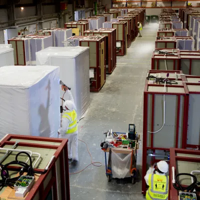 Factory workers preparing prefabricated construction modules inside a large manufacturing facility. Numerous partially assembled modules are arranged throughout the factory floor.