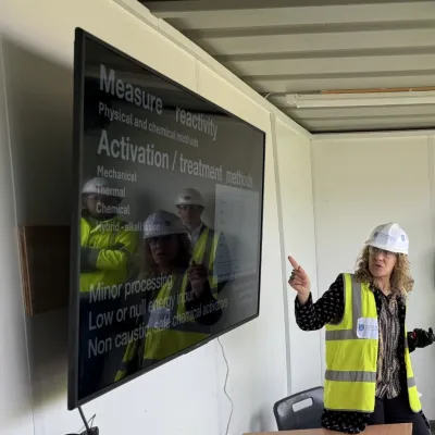 Woman in hard hat and safety vest presenting at a large screen display outdoors.