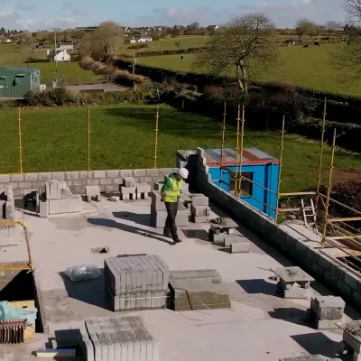 Construction worker in high-visibility clothing working on a building roof, surrounded by construction materials and scaffolding, with a rural landscape in the background.