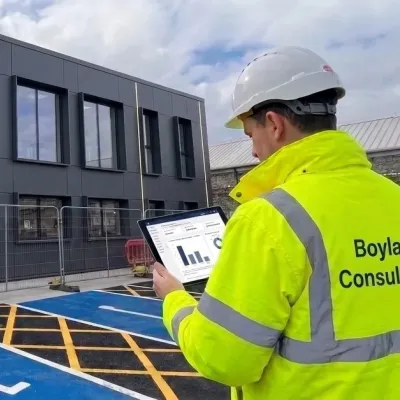Worker in high-visibility jacket reviewing documents beside a blue disabled parking bay at a modern building.