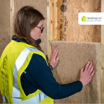 Woman in yellow safety vest installing mineral wool insulation panels in a wooden wall frame.