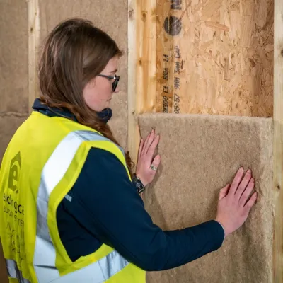 Worker in yellow safety vest installing insulation batts between wooden wall studs.