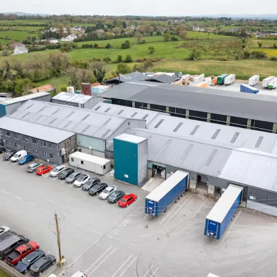 Aerial view of a large industrial warehouse complex with trucks and vehicles parked outside, surrounded by green countryside.