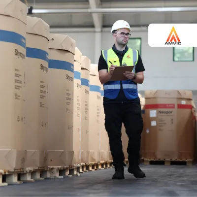 Warehouse worker in safety vest and hard hat checking inventory on tablet among large cardboard boxes.