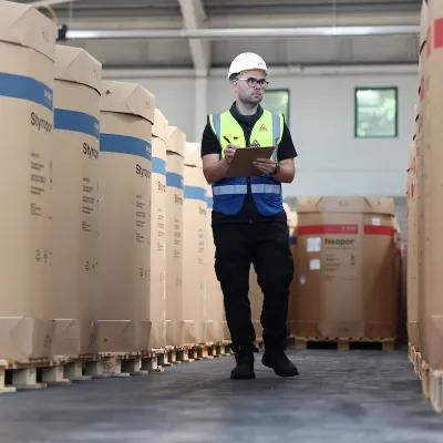 Worker in hard hat and safety vest walking through warehouse aisle with large cardboard boxes.