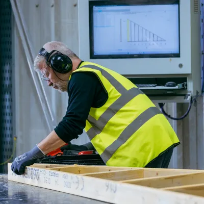 Worker in yellow safety vest and ear protection assembling wooden panels in a warehouse.