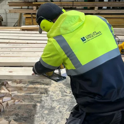 Worker in a yellow high-visibility jacket cutting timber planks at a lumber yard.