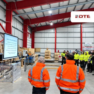 Two workers in orange hi-vis jackets facing a presentation screen in a large industrial warehouse, with a group of staff in yellow vests.