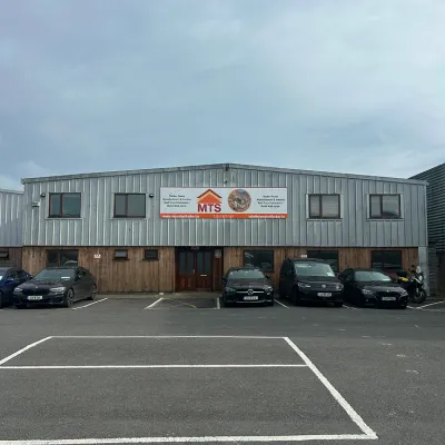 Industrial warehouse building with parked cars in front, under an overcast sky.