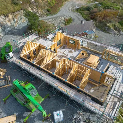 Aerial view of a wooden house frame under construction with green crane on a rocky hillside site.