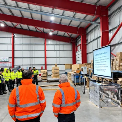 Group of workers in orange high-visibility vests attending a presentation inside a large warehouse.